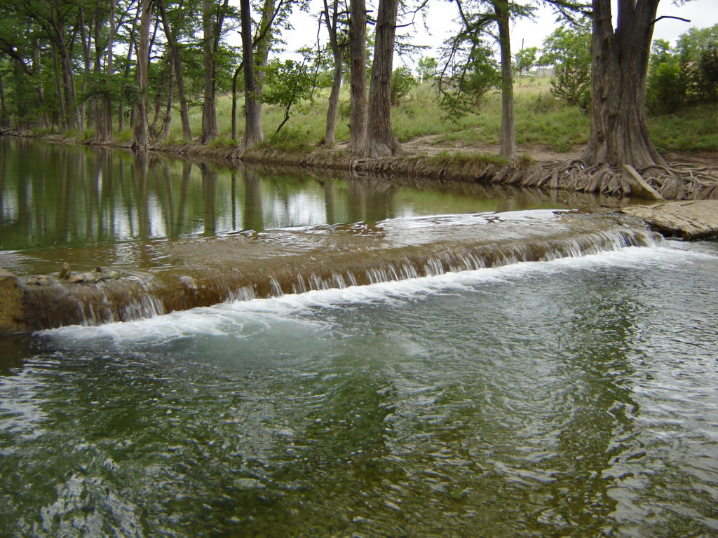 water flows over a small dam in a river with tree-lined banks