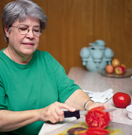 A senior woman with gray hair and green shirt is cutting tomatoes