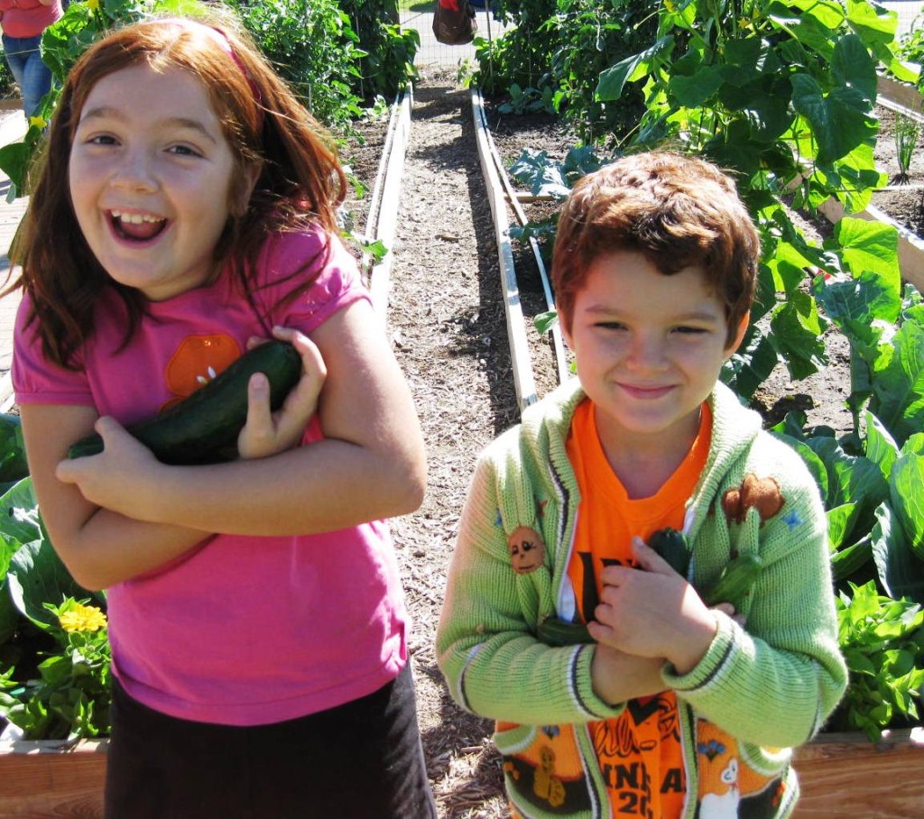 Young children holding vegetables they have grown. A red-headed girl holds an eggplant and an auburn-haired boy holds a squash. 