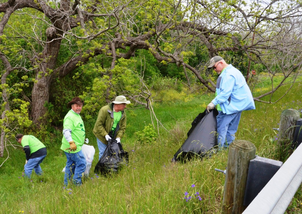 A group of volunteers in matching bright green T-shirts with trash bags are on a grassy embankment next to a guardrail. They pick up trash as part of the Geronimo and Alligator Watershed Cleanup Event.