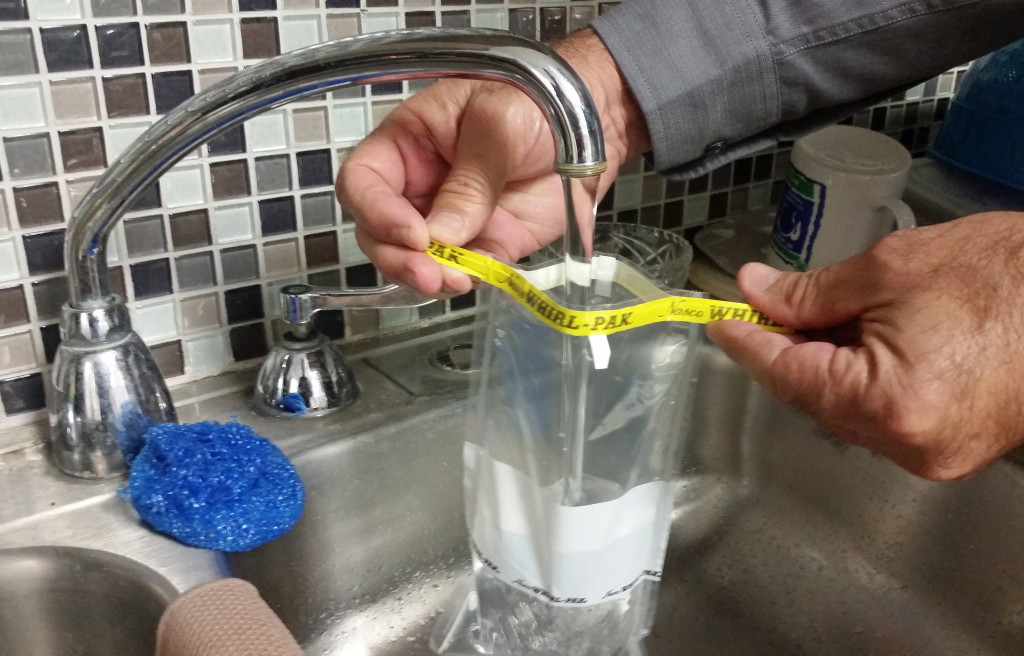 A water well screening clear bag is held underneath a tap. A man's hands hold the half-filled bag open under the running faucet