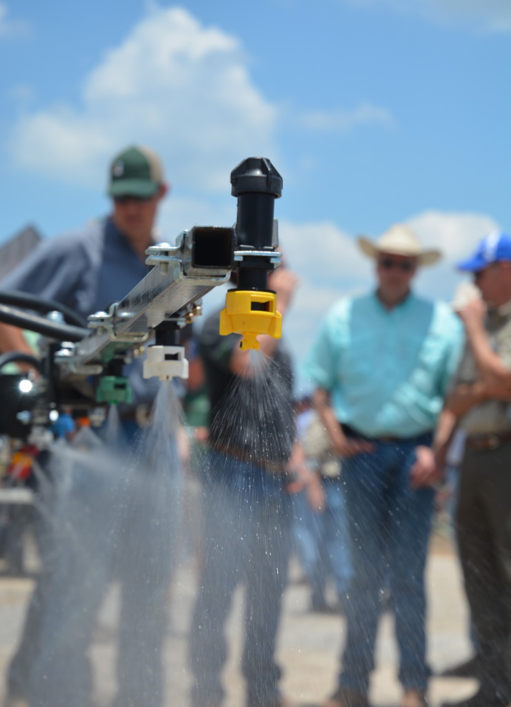 Applicator spraying pesticide in foreground, men in caps and cowboy hats are standing in the background against a blue sky.