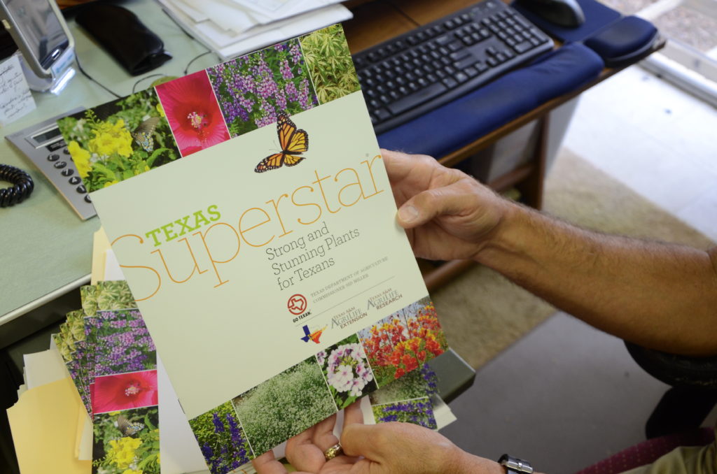 A man's hands hold a Texas Superstar plant guide that has come out of a printer on his desk. 