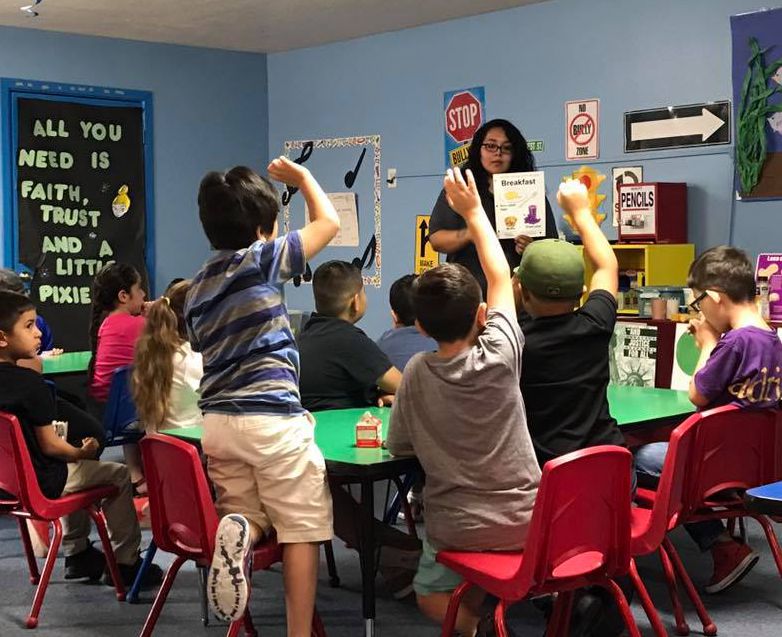 a group of young students raise their hand in a classroom setting - their backs are to us and the teacher looks on in back of the photo.