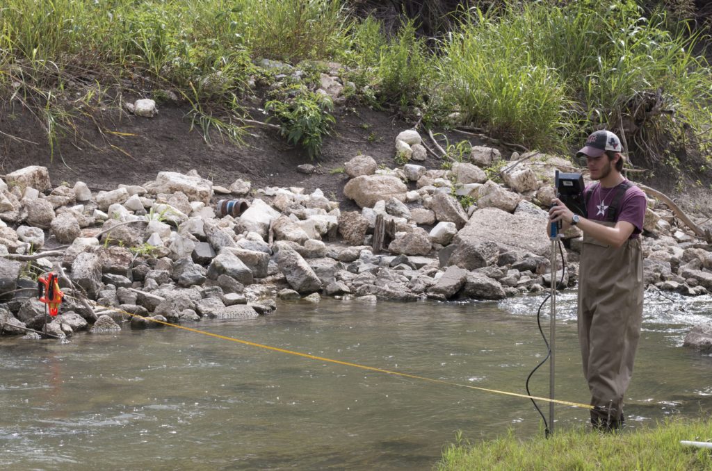 A man is monitoring Big Elm Creek. He wears waders and stands at the edge of the creek holding a machine to monitor water quality which can be impacted by area septic systems. 