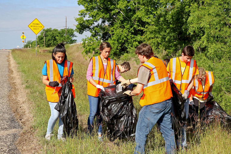 AgriLife photo of annual spring stream watershed cleanup. A group of people with trash bags wearing yellow reflective vests pick up debris by the side of a road.