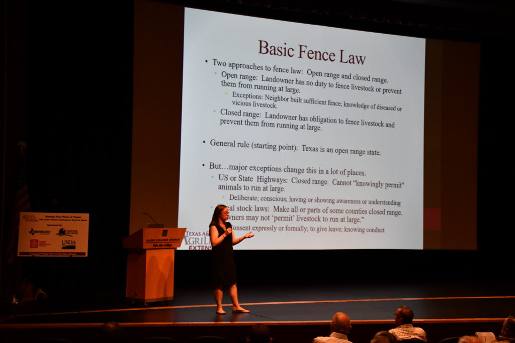 A woman in a dark dress and glasses stands in front of a large display screen in an auditorium. The title of the screen is Basic Fence Law.