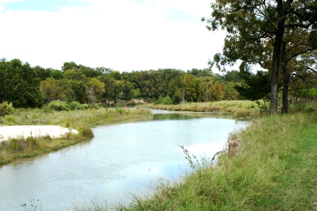 The Llano River in Texas. The water is quiet with green grasses, trees and woodland areas on either side of the banks.