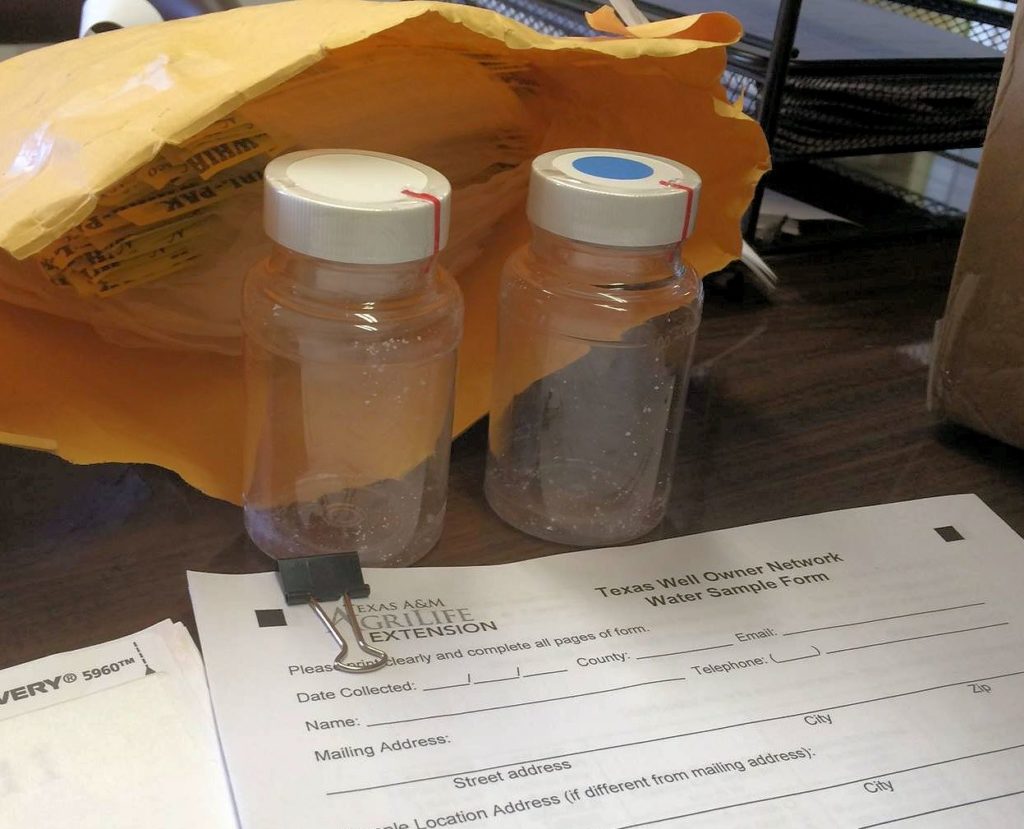Two small clear plastic vials to be used for water well sampling sit on a desk in front of an open, padded mailing envelope with testing forms on the desk in front of them.