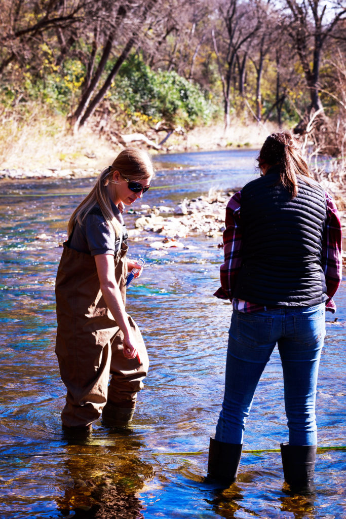Two young women stand in a stream. One wears waders while the other had high rubber boots on. One carries water quality testing equipment.