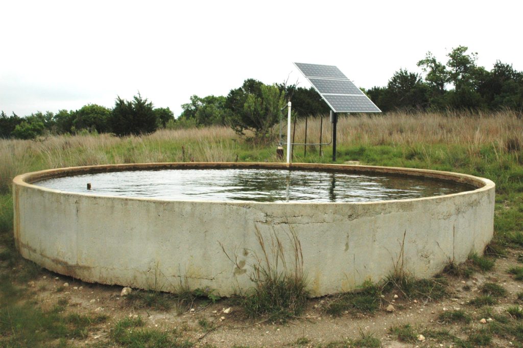 A large cement tank filled with well water. A solar panel sits next to it in a pasture.