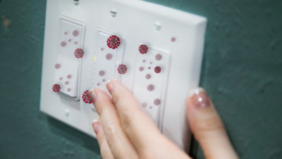 Female hand reaches to turn off a light switch with COVID-19 virus on the surface.
