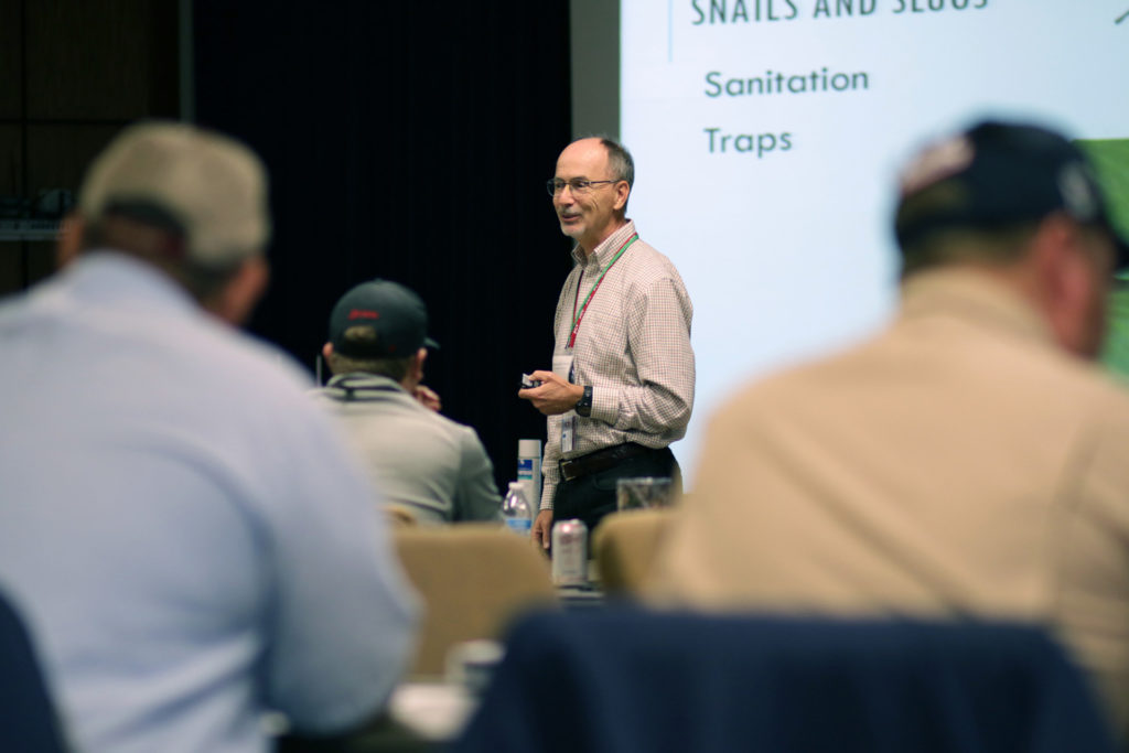 Man, Michael Merchant, standing at the front of a classroom teaching pest control course in school auditorium