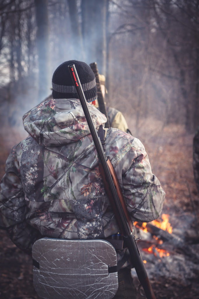 campfire burns behind two men dressed in hunting gear with guns