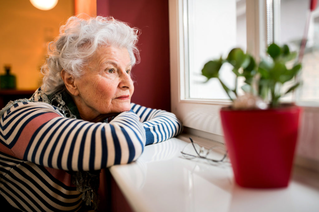 Sad alone senior woman looking through window at home, loneliness concept