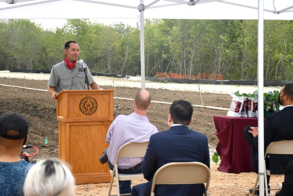 Man standing at a podium with people under a tent and a field at Greenies in the background