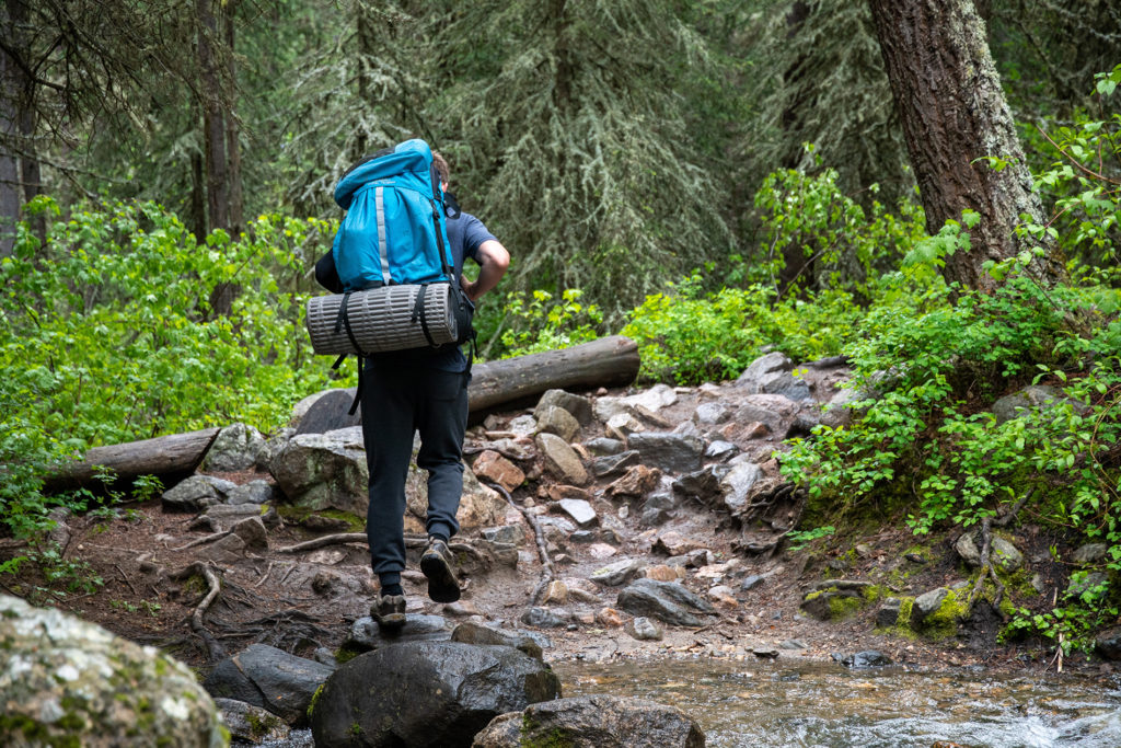 Man walking with a blue backpack across a stream and into a forested wilderness