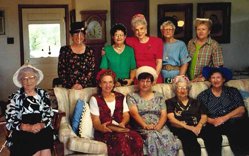 10 ladies sitting and standing, all with hats on, at a meeting in June Helwig's home setting