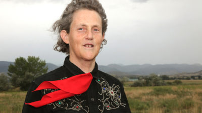 Temple Grandin standing outdoors, her hair and red neckerchief blow in the wind