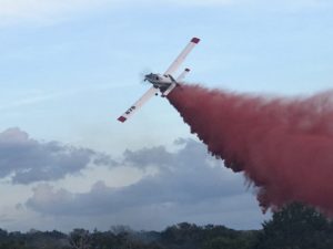A prop plane flies through the air releasing a red plume of fire retardant