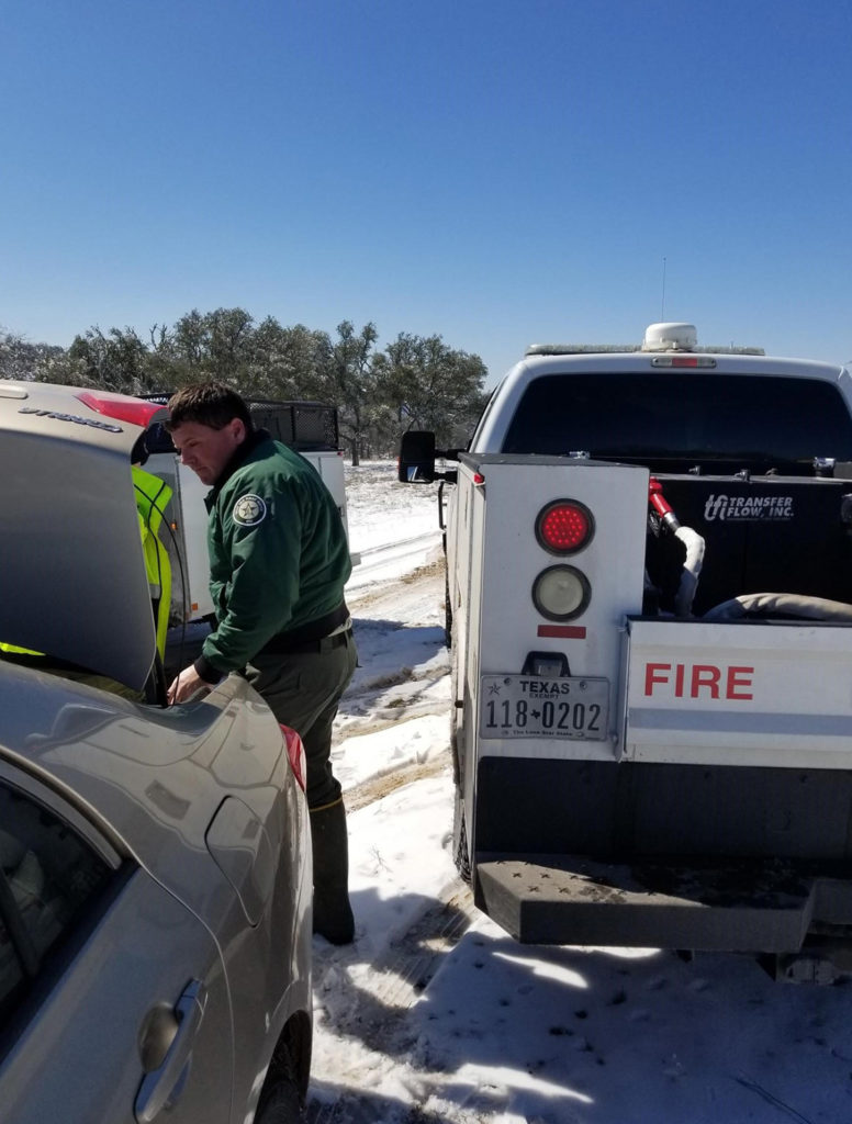 Heroic Texas A&M forester in uniform puts supplies into trunk of automobile. Four-wheel drive pickup trucks are in the background.