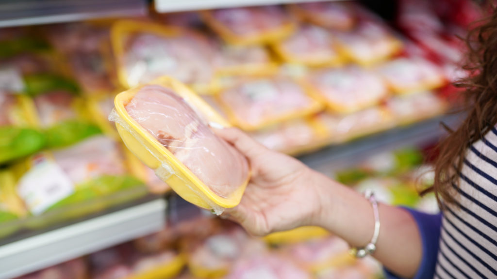 A close up of a woman's hand holding a package of raw chicken breasts in a supermarket aisle.