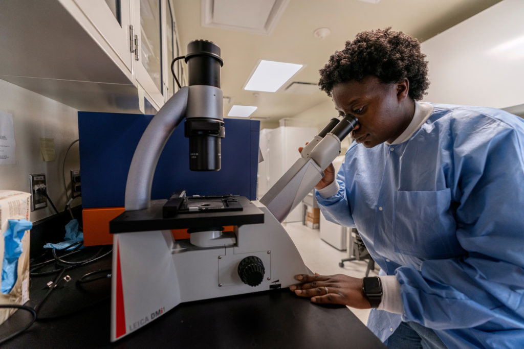 A Black person sits at a microscope in a lab setting