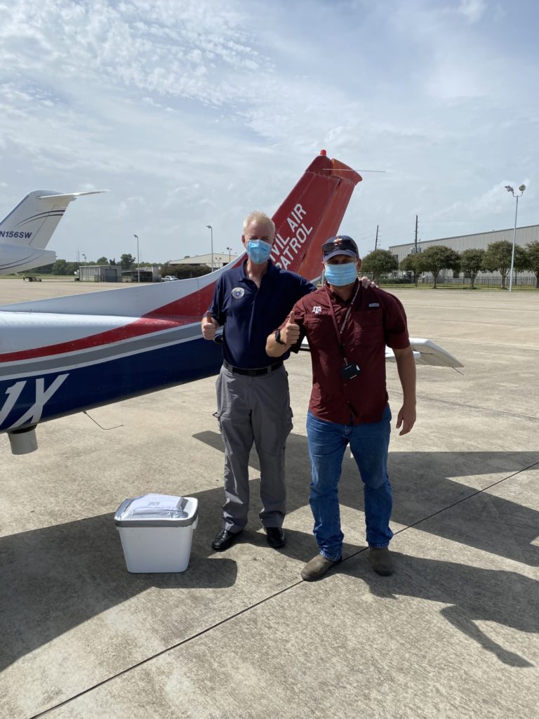 Two masked men standing by the tail of a small airplane that says Civil Air Patrol with a white tote at their feet - DAR agent making COVID test delivery to Civil Air Patrol pilot.