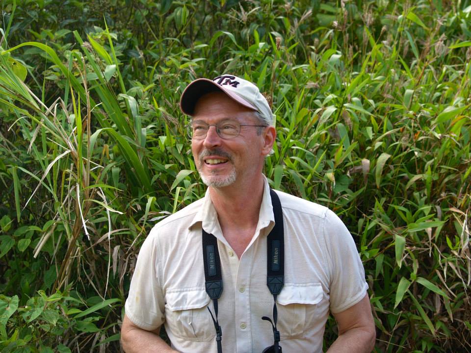 Photo of a white man with glasses and a grey/white mustache/goatee. He is wearing a white collared shirt, has a camera around his neck, and is wearing a white Texas A&M hat. He is standing in front of lush green tropical plant life. Lacher retires at the end of June 2021.