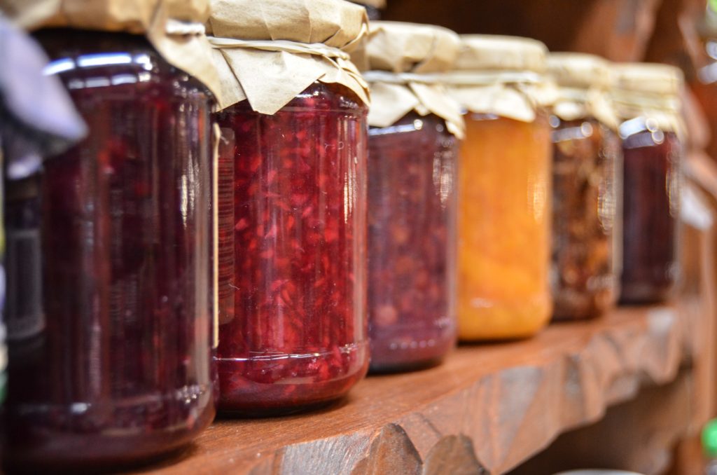 Canned food in mason jars on a shelf. An assortment of fruits and jellies in reds and yellows.