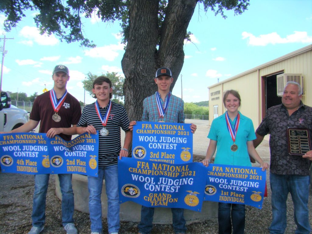 Four students hold blue signs, wear medals and stand beside their coach. The signs indicate they are the 2021 Grand Champion FFA wool judging team and their individual placings.