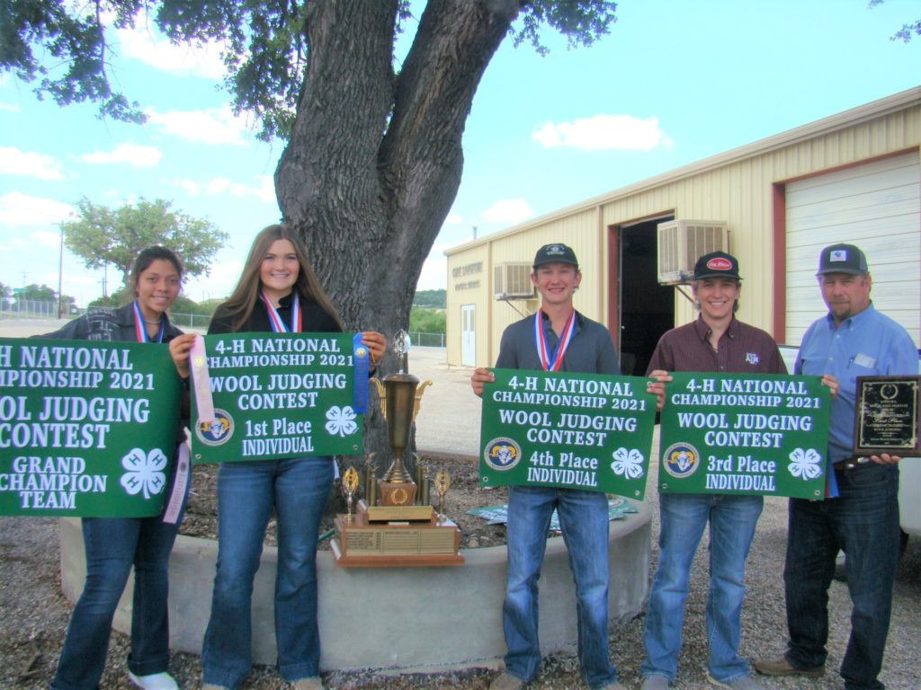 Four students holding green signs and wearing medals stand beside their coach under a tree. The signs indicate they are the 2021 champion 4-H wool judging team along with their individual placings.