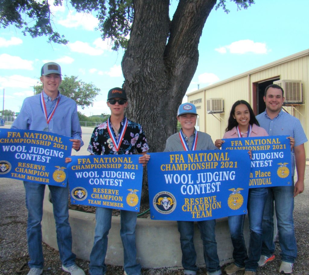 Four students hold blue signs and wear medals standing beside their male coach. The signs indicated they are the 2021 FFA reserve champion wool judging team and individual placings. 