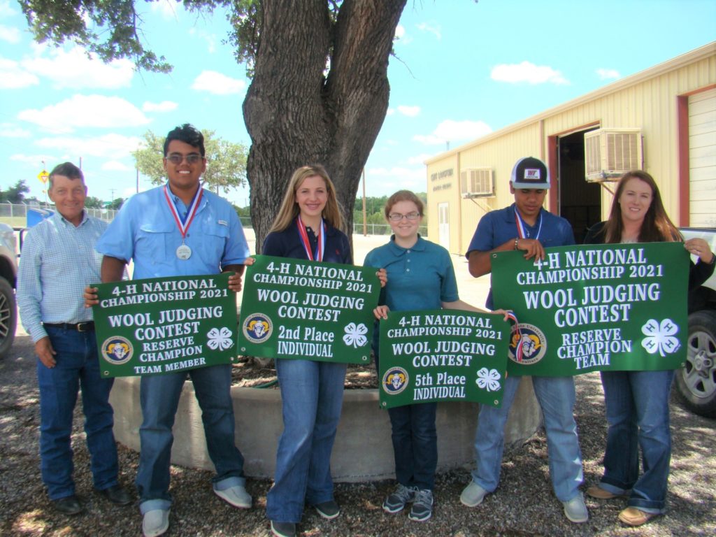 Five students hold green signs and wear their medals as they stand beside their male coach under a tree. The signs recognize them as the 2021 4-H wool judging reserve champion. 