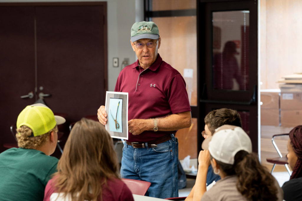 Man standing in a maroon shirt shows a picture to students who are sitting with the backs of their heads showing.