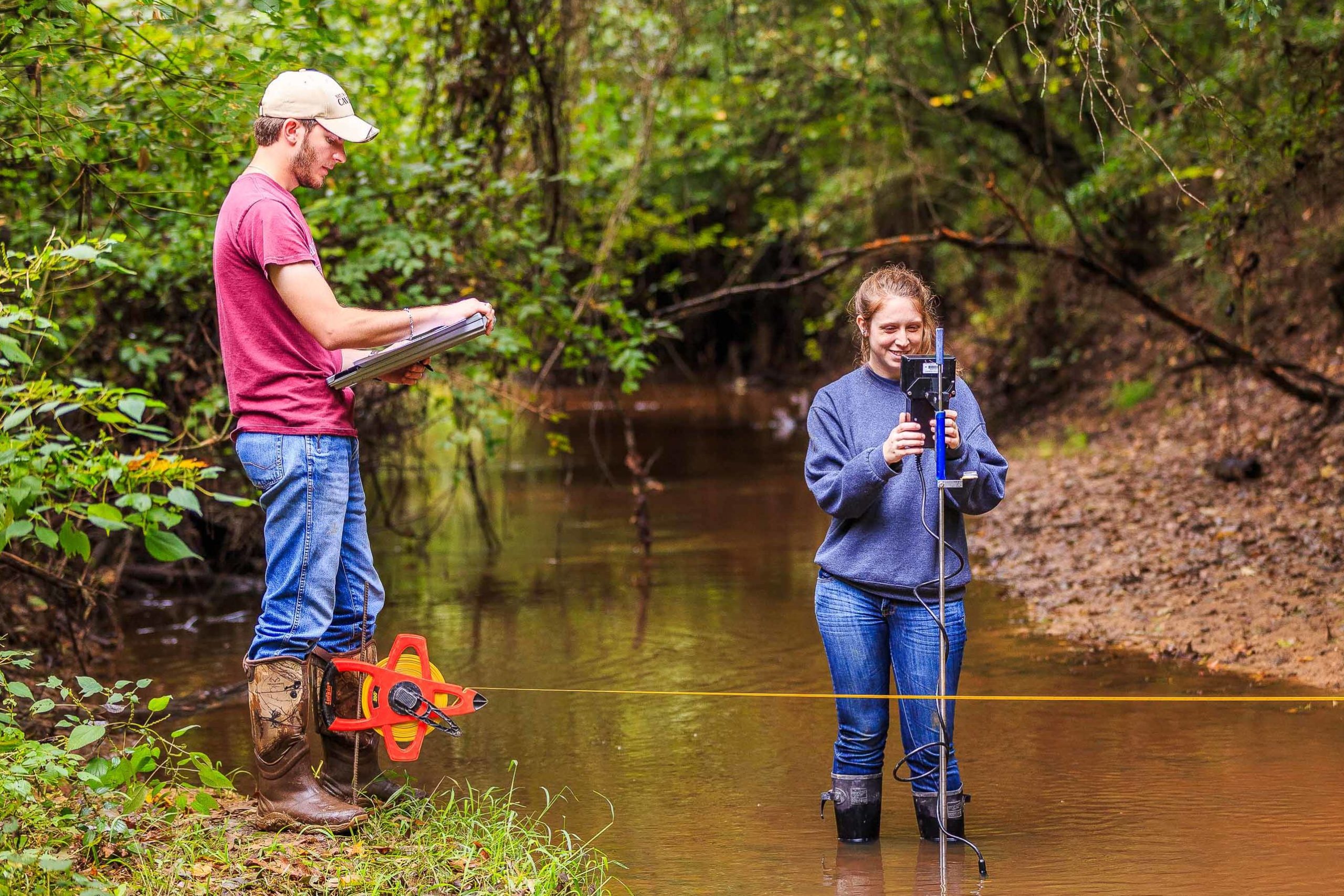 A woman wearing a blue sweatshirt, blue jeans, and knee-high waders stands in the middle of a shallow creek and is looking at a device that is attached to the top of a stick. This is Stephanie deVilleneuve. On the bank of the creek on the left side of the photo, a  man wearing a white baseball cap, red t-shirt, blue jeans and cowboy boots is taking notes on a clipboard. 