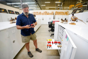 Man, Gary Voelker, standing in front of rows of white cabinets that hold the Collection of Birds, with one drawer pulled out and red and yellow birds laying on it.