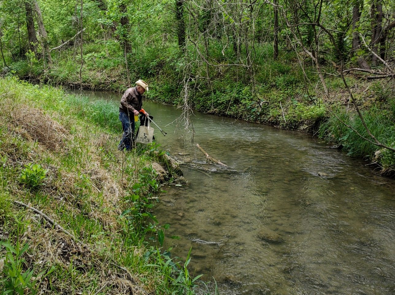 An older white man wearing a straw-colored cowboy hat, a long-sleeved shirt, and blue jeans stands on the bank of a shallow curving creek. He is holding a grabbing tool and a bucket. He appears to be reaching for something at the edge of the water with the grabbing tool. The man is a participant from the Spring Cleanup Event for the Geronimo and Alligator creek watershed.