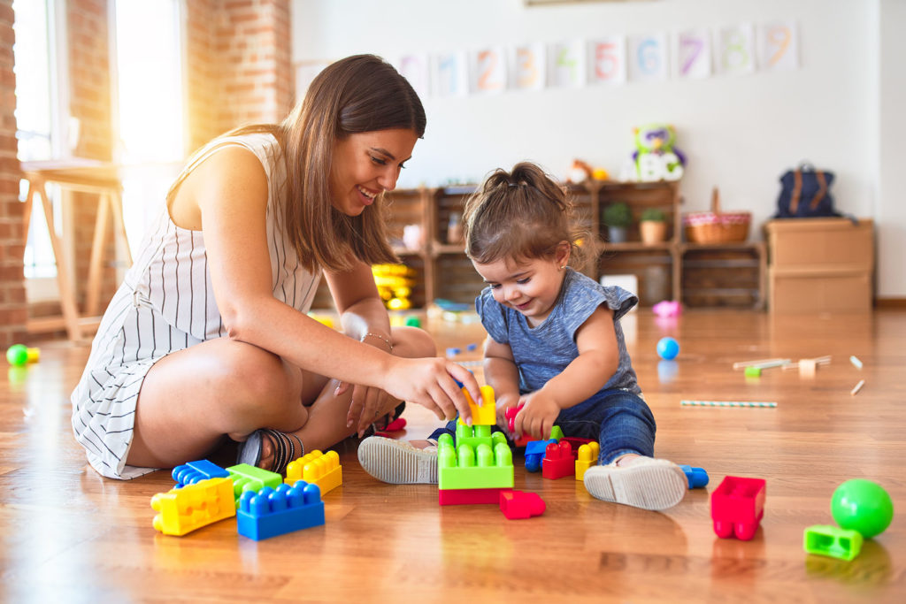 A woman providing child care with brown hair sits on the floor with a toddler with her hair in a ponytail and a blue outfit playing with blocks