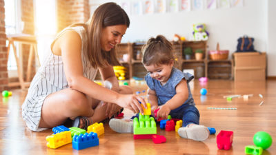 A woman sits on the floor with a toddler playing with blocks