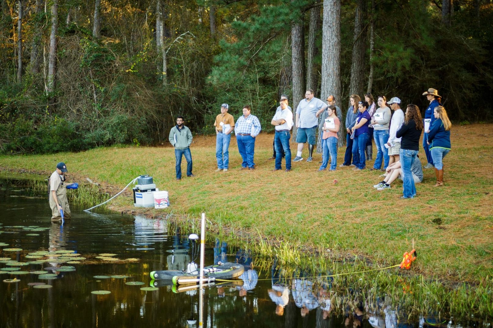 A group of people stand at the edge of a large pond. This is a photo of a past Watershed Planning short course.