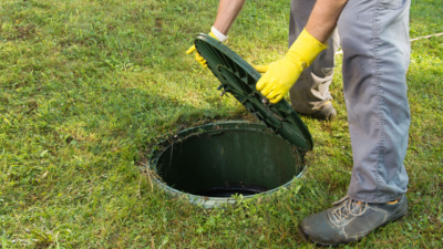 A man wearing yellow gloves holds open a septic tank lid in the middle of grass
