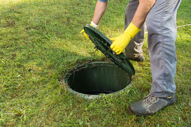A man wearing yellow gloves holds open a septic tank lid in the middle of grass