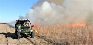 A green buggy drives down a field edge lighting a prescribed fire on dried grassland.
