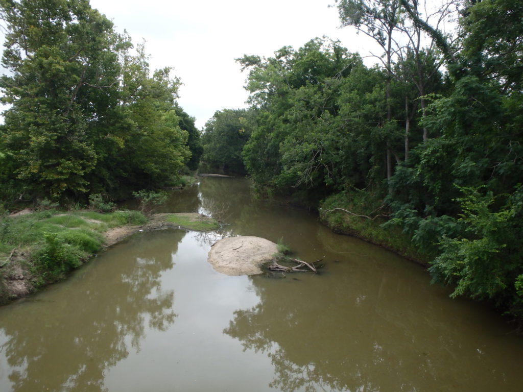 An image of a muddy, still creek lined on either side by green trees and foliage. The photo is elevated and taken from the perspective of the middle of the creek, suggesting the photographer was standing halfway across a bridge over the creek. There is a sandbar in the middle of the creek. This is the Mill Creek.