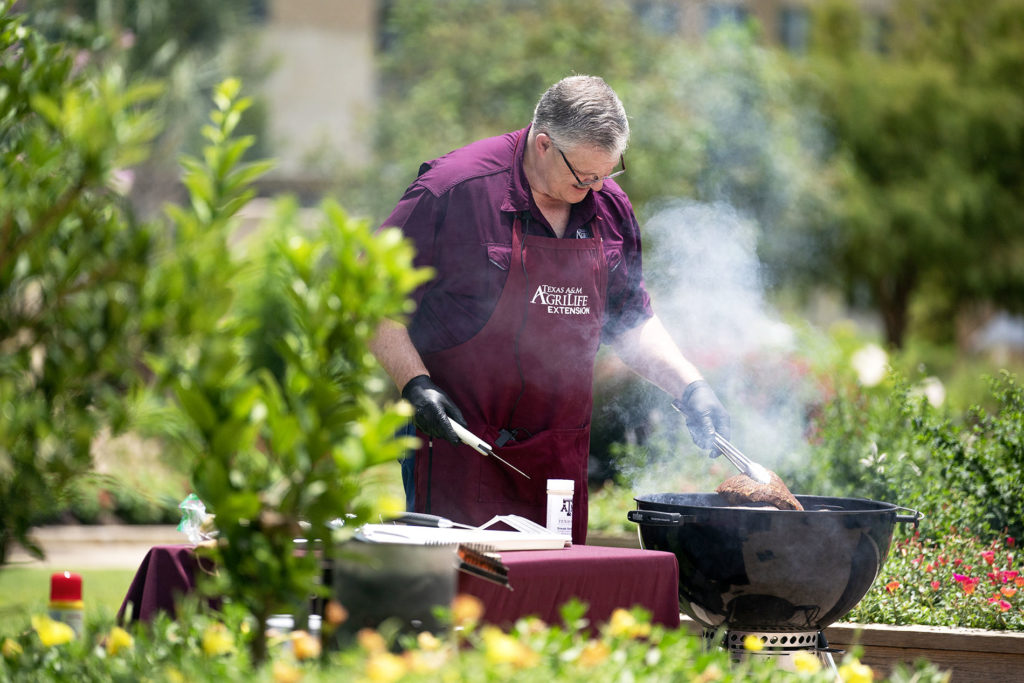 Davey Griffin - man dressed in a Texas A&M AgriLife Extension maroon apron - watches over food on a black grill that is sending up smoke.