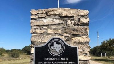 A historical marker at the Texas A&M AgriLife Research Sonora Station against a bright blue sky with an American flag flying