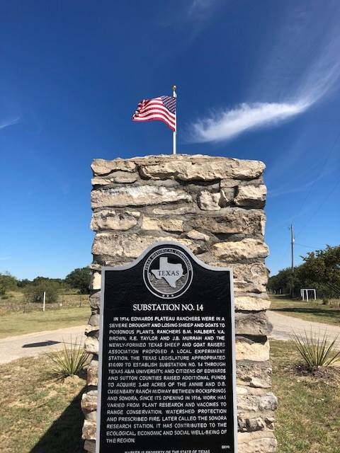 A historical marker at the Texas A&M AgriLife Research Sonora Station against a bright blue sky with an American flag flying