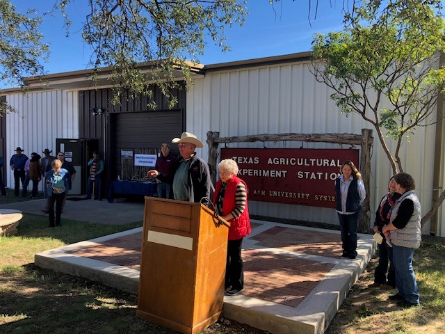A man and woman at a podium at the Sonora Station historical marker dedication. They stand outside in the sun with a metal building behind them 