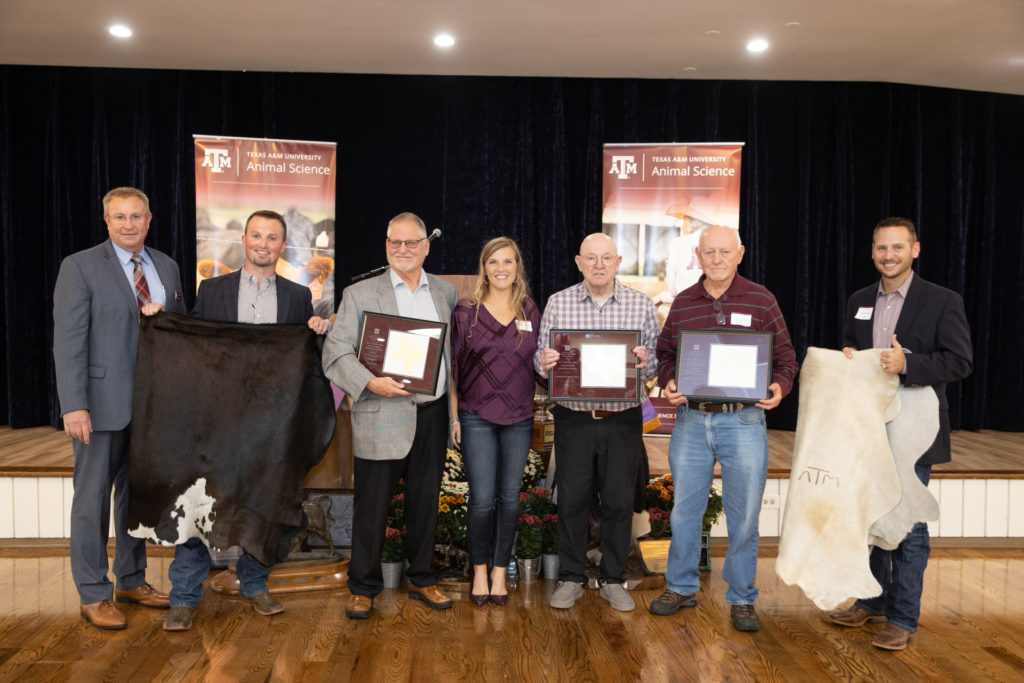 A group of people stand on a stage holding various plaques and cowhides, with banners behind them.